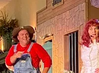 Two performers in costume, one in overalls and a hat and the other in a dress, are entertaining on a stage set resembling a rustic jail.