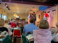 The image shows a live performance on stage in front of an audience seated at tables in a restaurant or theater setting.