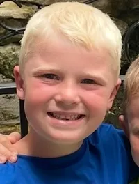 Two young boys wearing Batman shirts stand happily together in front of a small waterfall and greenery.