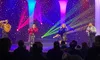 Performers dressed in sparkly outfits are singing on a colorful, illuminated stage in front of an audience.