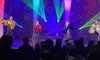 Four performers in glittery outfits are singing on a brightly lit stage with a colorful backdrop in front of an audience.