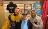 Three men are posing together, with two wearing outfits with shiny fringe and standing on either side of an older man in a blue shirt and cap, in front of a wall displaying ABBA and Beach Boys memorabilia.