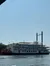 A large paddle steamboat with two tall smokestacks is floating on a calm body of water under a clear blue sky.