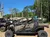 The image shows off-road vehicles with camo patterns parked on a gravel area surrounded by tall trees under a clear blue sky.