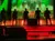 Five performers in sparkling jackets stand on a stage with vibrant red and green lighting.