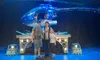 A group of three people is posing in front of a futuristic helicopter on a stage under blue lighting.