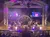 A musical performance is taking place on a decorative stage with dancers and musicians, under colorful lighting and digital backgrounds.