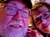 A smiling couple is taking a selfie in a room with a person blurred in the background.