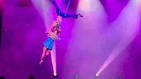 Two aerial performers are gracefully suspended in the air under vibrant purple stage lights.