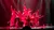 A group of performers in colorful costumes strike dramatic poses on a dimly lit stage under red lighting.