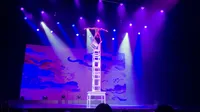A performer balances atop a tall stack of chairs under colorful stage lights.