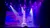 A performer balances atop a tall stack of chairs under colorful stage lights.