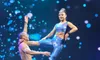 Two performers in sparkly blue outfits are balancing together on a platform, surrounded by a backdrop of blue lights.