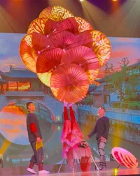 A performer balances multiple red umbrellas with their feet while supported by assistants on stage with a vibrant backdrop.