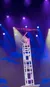 An acrobat performs a balancing act on a tower of chairs, illuminated by stage lights.