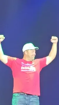 Two people are on stage, with one raising their arms triumphantly, while an audience watches.