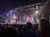 The image shows a crowded concert with vibrant stage lighting and a large audience watching performers.