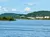 The image shows a scenic view of a lake with surrounding green hills and several residential buildings along the shore under a clear blue sky.