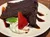 A slice of rich chocolate cake drizzled with chocolate sauce is served on a plate, accompanied by whipped cream, a strawberry, and a mint leaf.