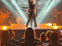 A performer strikes a dynamic pose on stage amidst bright lights and a rock band, with an audience watching intently.