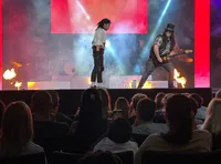 Two performers on stage, with one singing and the other playing guitar, are illuminated by colorful lights and surrounded by smoke effects in front of an audience.