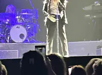 A performer on stage, wearing a sequined jacket and fedora, sings into a microphone while a crowd watches.
