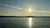 The image shows a serene sunset over a calm body of water with a silhouette of hills on the horizon, reflecting the sun's light.