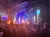 The image shows a live performance on stage with dramatic lighting and flames, viewed by an audience.