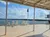 A covered outdoor area overlooks a body of water with scattered white chairs and a flag in the background under a partly cloudy sky.