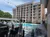 The image shows a multi-story hotel or apartment building with a pool in the outdoor area, surrounded by seating and ornate fencing, under a clear blue sky.