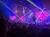 A performer is suspended upside down on a stage with dynamic lighting effects, while a large audience watches attentively.