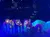 A vibrant stage performance features oversized, illuminated flowers and performers with colorful, fan-like props under blue lighting.