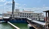 The image shows a large paddle boat docked at a pier on a sunny day, with clear skies and a calm body of water.