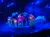 A vibrant stage set features oversized, glowing flowers and butterflies illuminated under blue stage lights.