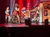 Four performers in matching red plaid skirts and black tops are energetically dancing on stage with a backdrop featuring rock band posters.