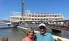 A couple is posing for a photo in front of a large paddlewheel steamboat docked on a sunny day.