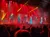 A group of performers in formal attire sing on a stage with festive red and yellow lighting effects before an audience.