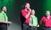 Four men on stage are smiling, dressed in red and green shirts with black suspenders, holding microphones.
