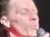 A person wearing a red shirt and black suspenders is speaking into a microphone on stage.