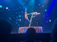 Two performers are balancing in an acrobatic pose on a circular platform with a blue, starry background.
