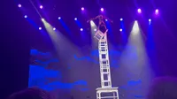 A performer balances on a tall, precarious stack of chairs under stage lights.