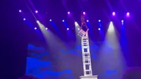 A performer is balanced upside down on a stack of chairs during a stage performance with dramatic lighting and a backdrop featuring blue cloud designs.