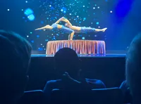 A contortionist performs a complex balancing act on a stage with a starry backdrop, watched by an audience.