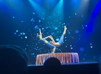 Two acrobats perform a complex balancing act on a stage with a blue, starry background and dramatic lighting.
