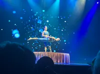 Two performers are on stage with one balancing horizontally on the other's head, surrounded by a backdrop of blue lights.