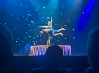 A duo performs an acrobatic routine on stage, with one person balancing the other on a small raised platform against a backdrop of blue lights.