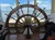 The image shows the interior of a boat with a large wooden ship's wheel and a view of the calm water outside through the windows.