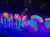 The image shows a colorful stage performance featuring large, illuminated flowers and butterflies in a dimly lit theater with an audience.