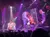 A group of musicians is performing energetically on stage with a harp under dramatic lighting and a large screen displaying their actions.