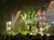 Performers on a stage are surrounded by vibrant yellow lighting and scaffolding, with a large screen displaying visuals in the background.